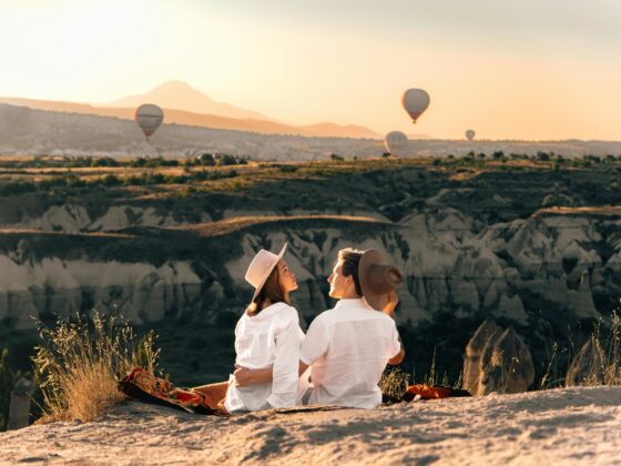 woman and man both wear white and sit on top of a cliff on a blanket. mountain view on the horizon as hot air balloons rise in the distance