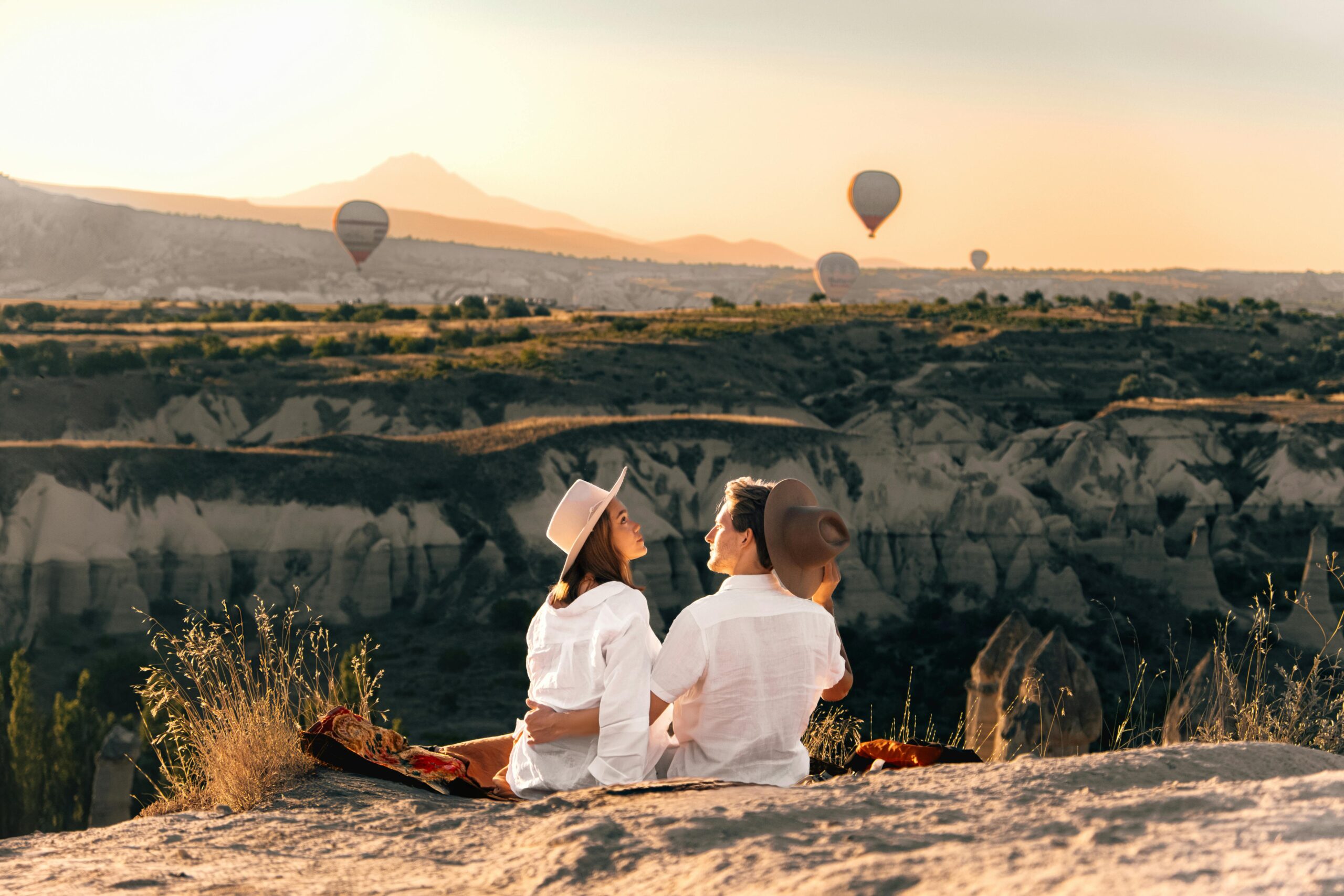 woman and man both wear white and sit on top of a cliff on a blanket. mountain view on the horizon as hot air balloons rise in the distance