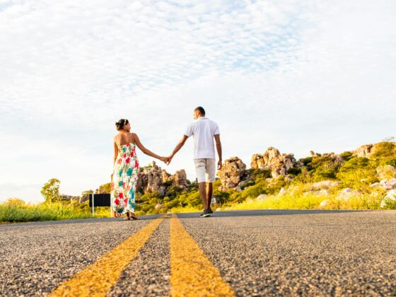 couple holding hands in distance as they walk down a street into the horizon with trees and blue sky