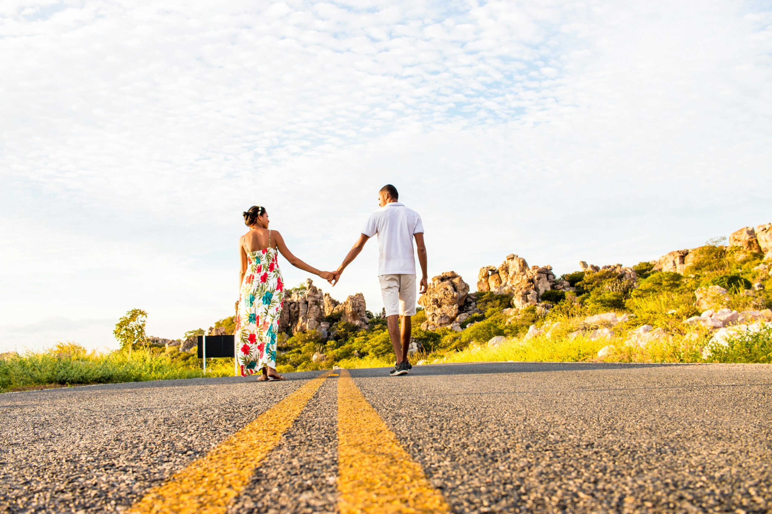 couple holding hands in distance as they walk down a street into the horizon with trees and blue sky