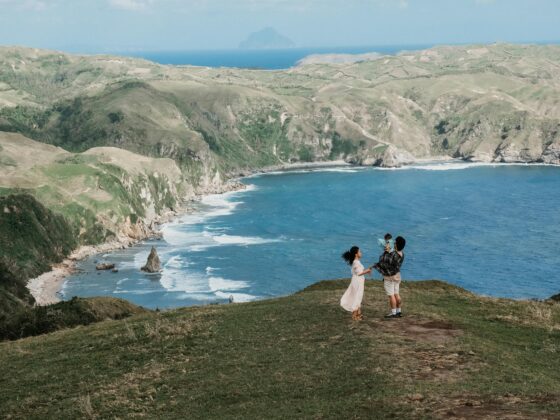 a mother father and young toddler stand together as father lifts boy up. they're on a cliff and rolling hills that overlook the water