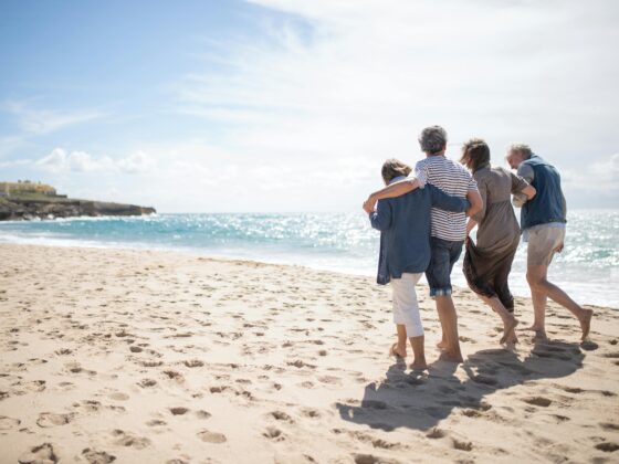 people walk arm in arm in the sand along the beach shoreline toward the horizon as the sun shines in a blue sky