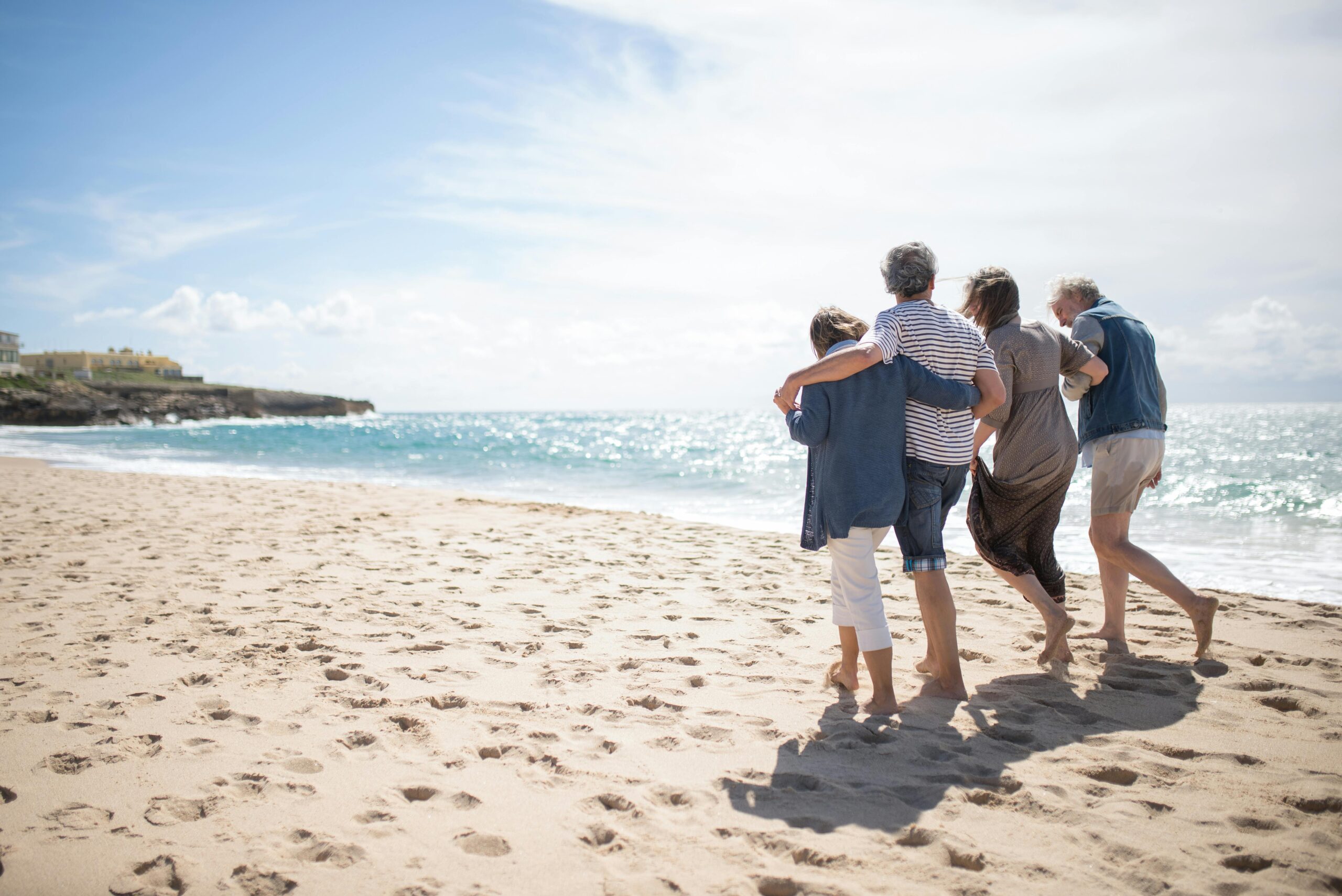 people walk arm in arm in the sand along the beach shoreline toward the horizon as the sun shines in a blue sky