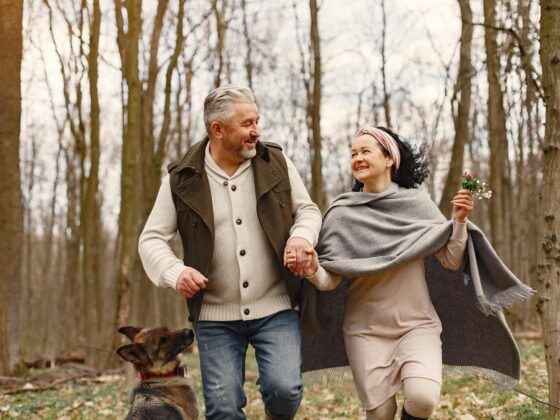 an older couple hold hands and look at each other smiling while walking through the woods with bare trees. a german shepard stands behind them and the woman holds a small bouquet of wild flowers