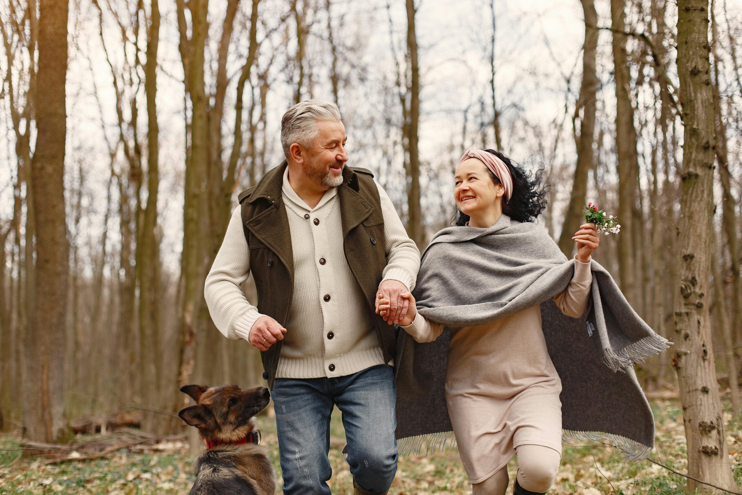 an older couple hold hands and look at each other smiling while walking through the woods with bare trees. a german shepard stands behind them and the woman holds a small bouquet of wild flowers