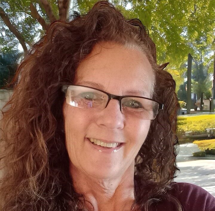 brunette woman smiles wearing glasses and a necklace. trees are behind her
